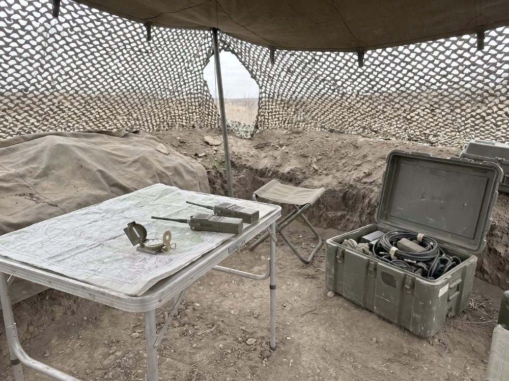 A field command post under camouflage netting, a map and field radios on the table beside an open equipment case